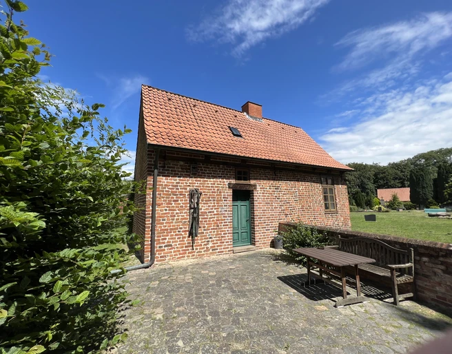 Kapitelhaus Wittlohe Rotes Ziegelhaus mit rotem Satteldach und grüner Tür, umgeben von einem gepflasterten Hof und Bäumen.