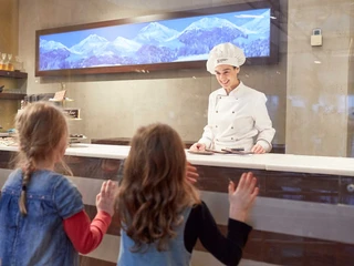 Chocolate Museum Eine Chocolatierin in weißer Uniform, umgeben von modernem Interieur, spricht mit drei Kindern.A chocolatier in a white uniform, surrounded by a modern interior, talks to three children.