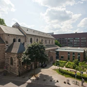 Museum Schnütgen Auf dem Bild ist das Museum Schnütgen in Köln zu sehen. Links erstreckt sich die romanische Cäcilienkirche, die Bruchsteinmauern und Rundbogenfenster zeigen. Rechts grenzt ein moderner Anbau an. Im Vordergrund ist ein kleiner Platz mit Bäumen und einigen Fahrrädern erkennbar.The picture shows the Schnütgen Museum in Cologne. On the left is the Romanesque Church of St. Cecilia, with its quarry stone walls and arched windows. On the right is a modern extension. A small square with trees and a few bicycles can be seen in the foreground.