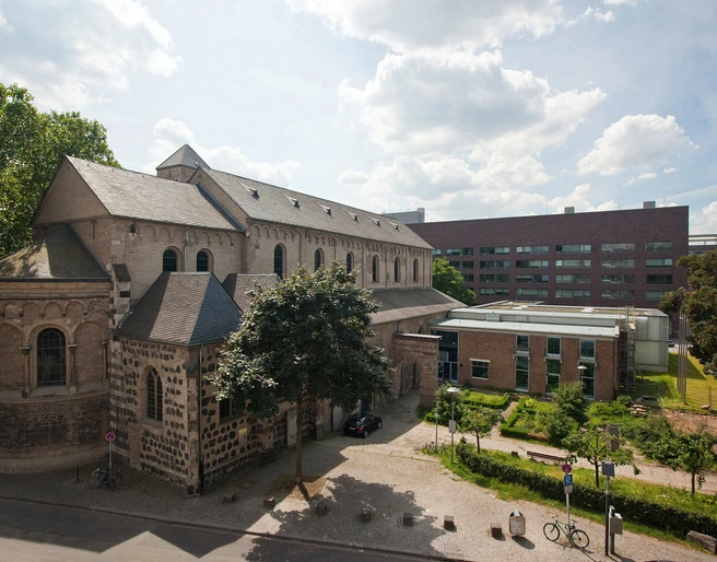 Museum Schnütgen Auf dem Bild ist das Museum Schnütgen in Köln zu sehen. Links erstreckt sich die romanische Cäcilienkirche, die Bruchsteinmauern und Rundbogenfenster zeigen. Rechts grenzt ein moderner Anbau an. Im Vordergrund ist ein kleiner Platz mit Bäumen und einigen Fahrrädern erkennbar.The picture shows the Schnütgen Museum in Cologne. On the left is the Romanesque Church of St. Cecilia, with its quarry stone walls and arched windows. On the right is a modern extension. A small square with trees and a few bicycles can be seen in the foreground.