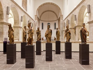 Museum Schnütgen Das Bild zeigt einen hell erleuchteten Museumsraum mit einer Reihe von hölzernen Skulpturen auf Sockeln.The picture shows a brightly lit museum room with a series of wooden sculptures on pedestals.