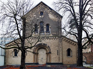 St. Cecilia Das Bild zeigt die romanische Kirche Sankt Cäcilien in Köln. Die schlichte Backsteinfassade wird von zwei Bäumen flankiert, deren kahle Äste die Sicht auf das Bauwerk umrahmen. The picture shows the Romanesque church of St. Cäcilien in Cologne. The simple brick façade is flanked by two trees whose bare branches frame the view of the building.