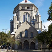 Saint Gereon Romanische Kirche aus Sandstein mit imposanter Kuppel und Buntglasfenstern, umgeben von Bäumen.Romanesque church made of sandstone with an imposing dome and stained glass windows, surrounded by trees.
