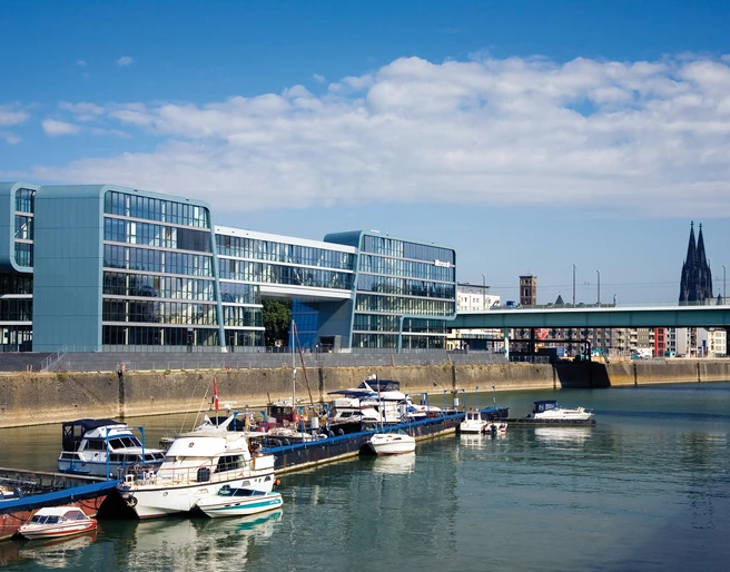 Rheinauhafen Modernes Bürogebäude am Wasser mit Booten im Vordergrund, blauer Himmel und Kölner Dom im Hintergrund.Modern office building on the waterfront with boats in the foreground, blue sky and Cologne Cathedral in the background.