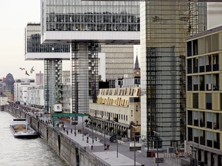 Rheinauhafen Auf dem Bild sind die Kranhäuser des Rheinauhafens in Köln zu sehen, die entlang des Rheinufers stehen. Es handelt sich um moderne, stufige Hochhäuser, die sich durch ihre architektonische Einzigartigkeit hervorheben. Im Vordergrund verläuft eine Promenade mit Menschen, während im Hintergrund weitere Gebäude und der Fluss sichtbar sind.The picture shows the crane buildings of the Rheinauhafen in Cologne, which stand along the banks of the Rhine. These are modern, stepped high-rise buildings that stand out for their architectural uniqueness. A promenade with people runs in the foreground, while other buildings and the river are visible in the background.