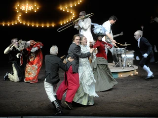 Schauspiel Ensemble in historischen Kostümen auf einer Bühne; Musiker und Schauspieler interagieren dynamisch.Ensemble in historical costumes on a stage; musicians and actors interact dynamically.