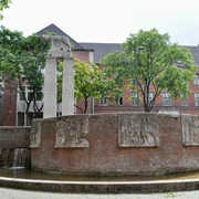Roman fountain Eine ovale Brunnenanlage mit reliefverzierten, rötlichen Backsteinwänden, flankiert von Bäumen und einem Gebäude mit roten Ziegelwänden im Hintergrund.An oval fountain with reddish brick walls decorated in relief, flanked by trees and a building with red brick walls in the background.