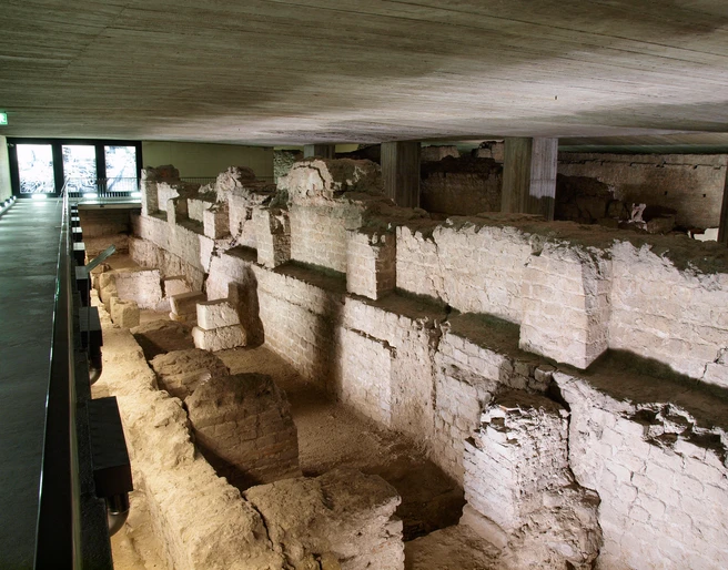 MiQua Das Bild zeigt die Überreste des römischen Statthalterpalastes, des Praetoriums, in Köln. Die alten Mauerstrukturen sind teilweise erhalten und durch schummriges Licht ausgeleuchtet, das die historische Atmosphäre unterstreicht.The picture shows the remains of the Roman governor's palace, the Praetorium, in Cologne. The old wall structures are partially preserved and illuminated by dim light, which emphasizes the historical atmosphere.