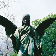 Melaten Cemetery Die majestätische Statue eines Engels mit weit geöffneten Flügeln erhebt sich erhaben vor einem Hintergrund aus blühendem Frühlingslaub und einem klaren Himmel.The majestic statue of an angel with wide-open wings rises majestically against a backdrop of blossoming spring foliage and a clear sky.