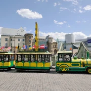 Wolters Bimmelbahn Eine gelb-grüne Bimmelbahn fährt über Kopfsteinpflaster, im Hintergrund moderne und historische Gebäude.A yellow and green train runs over cobblestones, with modern and historic buildings in the background.