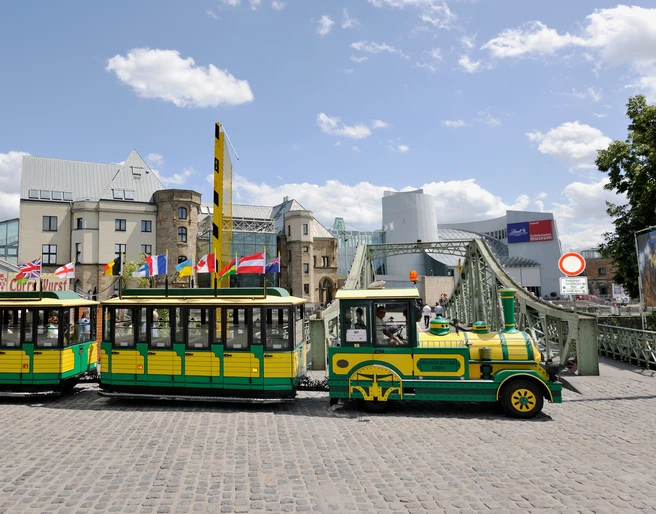 Wolters Bimmelbahn Eine gelb-grüne Bimmelbahn fährt über Kopfsteinpflaster, im Hintergrund moderne und historische Gebäude.A yellow and green train runs over cobblestones, with modern and historic buildings in the background.