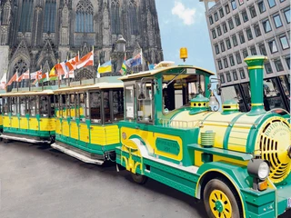 Wolters Bimmelbahn Eine gelb-grüne Bimmelbahn steht vor dem imposanten Kölner Dom, flankiert von einem modernen Gebäude.A yellow and green train stands in front of the imposing Cologne Cathedral, flanked by a modern building.