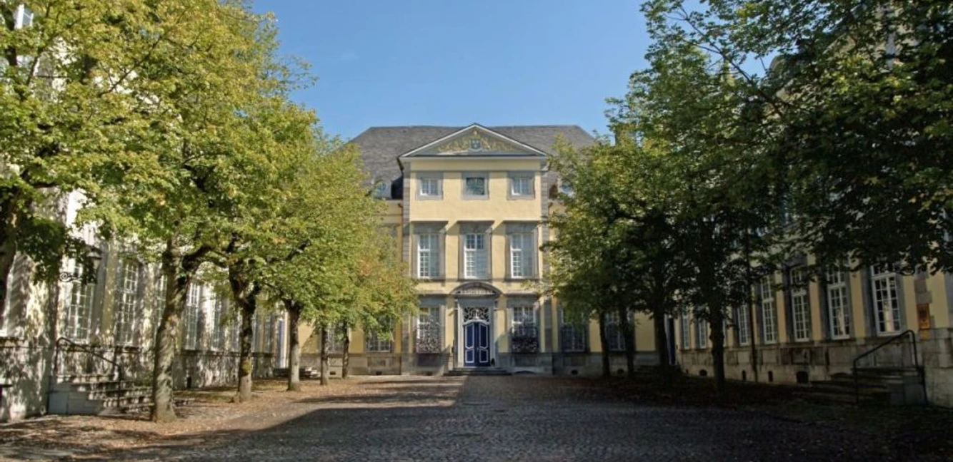 Reichsabtei Kornelimünster Historisches Gebäude mit gepflastertem Vorplatz zwischen zwei Baumreihen unter blauem Himmel.