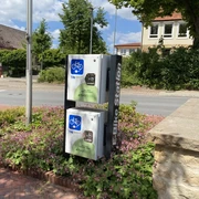 e_bike-ladestation_rathaus_cc_gemeinde-hagen-atw Ladestation für E-Bikes an einer Straßenkreuzung in einer grünen Umgebung.