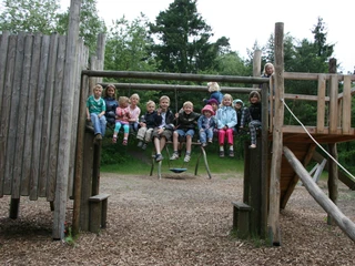 kinder-abenteuerspielplatz Eine fröhliche Gruppe von Kindern sitzt auf der oberen Plattform eines Abenteuerspielplatz-Klettergerüsts aus Holz, vor einem bewaldeten Hintergrund.