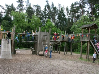 abenteuerspielplatz-luhegrund Ein großer Holzspielplatz mit Rutschen, Kletterstrukturen und spielenden Kindern, umgeben von Bäumen.