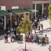 kleiner-überblick Ein belebter Platz mit modernen Geschäften, Menschen flanieren zwischen Boutiquen, Bänken und jungen Bäumen.