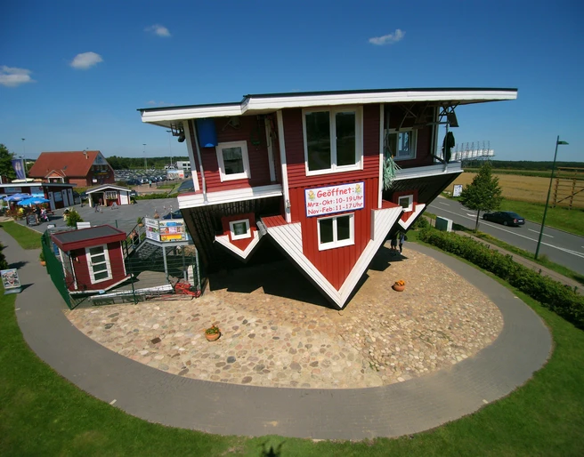 Das verrückte Haus Aussenansicht Das Bild zeigt ein auf dem Kopf stehendes, rot-weißes Holzhaus, umgeben von grünem Rasen und einer asphaltierten Straße.