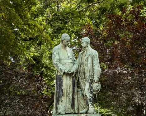 Adolf Kolping Monument Das Denkmal zeigt Adolf Kolping und einen jungen Mann in der typischen Arbeiterkleidung des 19. Jahrhunderts. Beide stehen im stilvollen Park, umgeben von dichtem Laub und einer Blumenrabatte. Das Denkmal ist auf einem Sockel montiert, auf dem der Name "ADOLF KOLPING" graviert ist.The monument shows Adolf Kolping and a young man in typical 19th century working-class clothing. Both are standing in the stylish park, surrounded by dense foliage and a flower border. The monument is mounted on a pedestal on which the name "ADOLF KOLPING" is engraved.