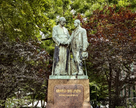 Adolf Kolping Monument Adolf Kolping Denkmal, bestehend aus zwei respektvoll gestalteten Bronzestatuen, umgeben von üppigen Bäumen.Adolf Kolping monument, consisting of two respectfully designed bronze statues, surrounded by lush trees.