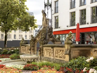 Pixies’ Fountain Eine üppige Blumengestaltung umgibt den Heinzelmännchenbrunnen mit seinen detailreichen Reliefs und Skulpturen, dahinter moderne Gebäude.A lush floral design surrounds the Heinzelmännchenbrunnen fountain with its detailed reliefs and sculptures, with modern buildings behind it.