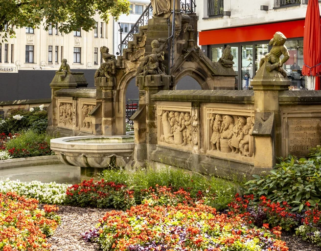 Pixies’ Fountain Heinzelmännchenbrunnen in Köln: Brunnen mit Figuren und Reliefs, umgeben von Blumen.Heinzelmännchenbrunnen in Cologne: fountain with figures and reliefs, surrounded by flowers.