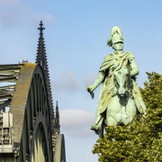 Equestrian statue of King Frederick William IV. Das Reiterstandbild König Friedrich Wilhelm IV. zeigt eine majestätische Bronzestatue auf einem Pferd. Im Hintergrund erhebt sich das Kölner Wahrzeichen, der Dom, während das Bild von der ikonischen Hohenzollernbrücke eingerahmt wird.The equestrian statue of King Frederick William IV shows a majestic bronze statue on a horse. Cologne's landmark, the cathedral, rises in the background, while the image is framed by the iconic Hohenzollern Bridge.
