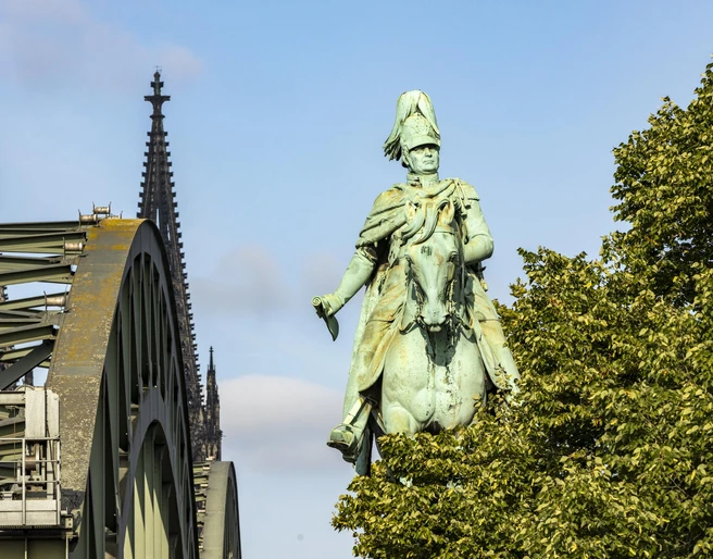 Equestrian statue of King Frederick William IV. Das Reiterstandbild König Friedrich Wilhelm IV. zeigt eine majestätische Bronzestatue auf einem Pferd. Im Hintergrund erhebt sich das Kölner Wahrzeichen, der Dom, während das Bild von der ikonischen Hohenzollernbrücke eingerahmt wird.The equestrian statue of King Frederick William IV shows a majestic bronze statue on a horse. Cologne's landmark, the cathedral, rises in the background, while the image is framed by the iconic Hohenzollern Bridge.