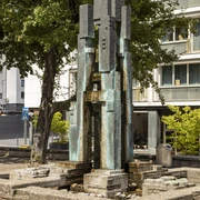 Theo Burauen Fountain Das Bild zeigt den Theo-Burauen-Brunnen, einen eleganten, vertikalen Steingussbrunnen in Köln. Umliegend sind ein grüner Baum und moderne Gebäude sichtbar, die das urbane Stadtbild ergänzen.The picture shows the Theo Burauen fountain, an elegant, vertical cast stone fountain in Cologne. A green tree and modern buildings are visible around it, complementing the urban cityscape.