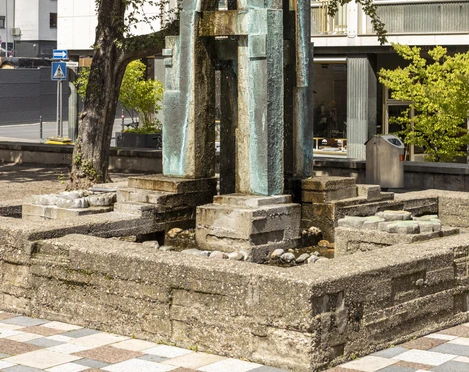 Theo Burauen Fountain Das Bild zeigt den Theo-Burauen-Brunnen, einen eleganten, vertikalen Steingussbrunnen in Köln. Umliegend sind ein grüner Baum und moderne Gebäude sichtbar, die das urbane Stadtbild ergänzen.The picture shows the Theo Burauen fountain, an elegant, vertical cast stone fountain in Cologne. A green tree and modern buildings are visible around it, complementing the urban cityscape.