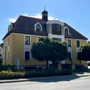 Touristinformation Dittersbach-(c)-emily-wolf-CC-BY-(1).jpg Gelbes zweistöckiges Gebäude mit rotem Dach und Turmuhr, umgeben von Bäumen und Sträuchern, bei sonnigem Wetter.
