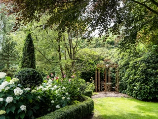 garten-landhof-tausendschoen-apen-5 Ein liebevoll gepflegter Garten mit farbenfrohen Blumenbeeten, einem grünen Rasen und einer eleganten Metalllaube, die von Rankgewächsen umgeben ist. Üppig blühende Hortensien im Vordergrund, dichter Baum- und Sträucherbestand im Hintergrund.