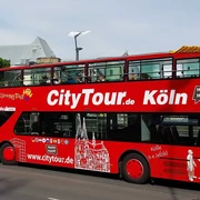 Cologne CityTour Ein roter Doppeldecker-Bus der „CityTour Köln“ steht am Straßenrand, mit geöffnetem Oberdeck. Die weißen Illustrationen von Kölner Wahrzeichen zieren den Bus.A red double-decker "CityTour Köln" bus is parked on the side of the road with its upper deck open. The bus is adorned with white illustrations of Cologne landmarks.