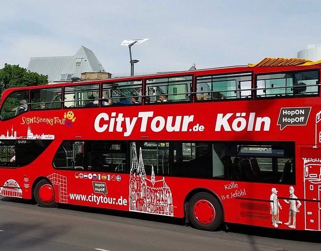 Cologne CityTour Ein roter Doppeldecker-Bus der „CityTour Köln“ steht am Straßenrand, mit geöffnetem Oberdeck. Die weißen Illustrationen von Kölner Wahrzeichen zieren den Bus.A red double-decker "CityTour Köln" bus is parked on the side of the road with its upper deck open. The bus is adorned with white illustrations of Cologne landmarks.