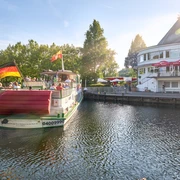 Wasserbahnhof, Mülheim an der Ruhr Ein Passagierschiff mit Deutschlandflagge an einem Flussanleger, flankiert von Bäumen und einem Gebäude.