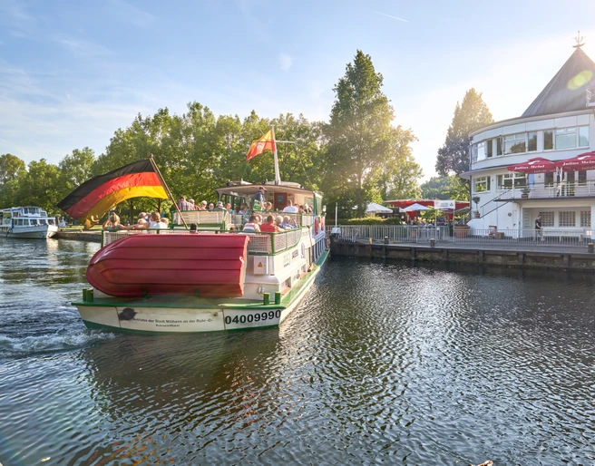 Wasserbahnhof, Mülheim an der Ruhr Ein Passagierschiff mit Deutschlandflagge an einem Flussanleger, flankiert von Bäumen und einem Gebäude.