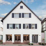 Hausansicht. Eine dreistöckige, weiße Hausfassade. Im Erdgeschoss drei große Fenster, darüber Fenster mit Fensterläden