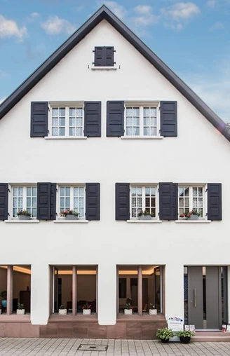 Hausansicht. Eine dreistöckige, weiße Hausfassade. Im Erdgeschoss drei große Fenster, darüber Fenster mit Fensterläden