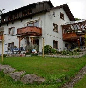 Dreistöckiges weißes Haus mit dunklem Dach. Mehrere Holzbalkone, Terrasse Schaukel. Abfallende Rasenfläsche