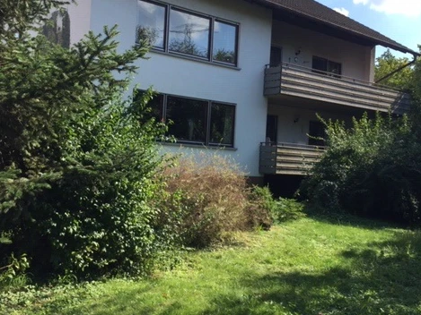 Außenansicht, weiße Hausfassade und zwei Balkone, davor Garten mit Büschen