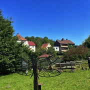 Eine Metallskulptur einer Libelle steht auf einer grünen Wiese, umgeben von Häusern und Bäumen.