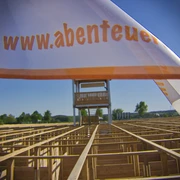 abenteuer-labyrinth-fahne Das Bild zeigt eine weiße Fahne mit der Aufschrift "www.abenteuer..." im Vordergrund. Im Hintergrund ist ein großes Labyrinth aus Holz zu sehen, über dem ein hoher Aussichtsturm steht.