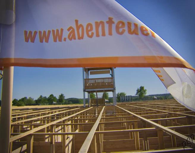 abenteuer-labyrinth-fahne Das Bild zeigt eine weiße Fahne mit der Aufschrift "www.abenteuer..." im Vordergrund. Im Hintergrund ist ein großes Labyrinth aus Holz zu sehen, über dem ein hoher Aussichtsturm steht.