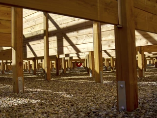labyrinth-bispinger-heide Das Bild zeigt die Ansicht unterhalb eines hölzernen Heckenlabyrinths in der Bispinger Heide mit Kieselsteinen am Boden, Sonnenlicht und Schatten werfen geometrische Muster auf die Holzkonstruktion.