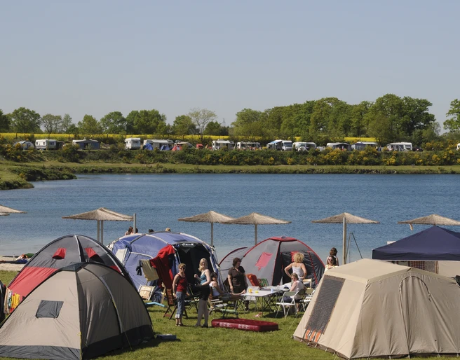 Am Ufer eines Sees stehen zahlreiche bunte Zelte. Menschen sitzen entspannt zusammen.