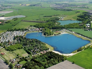Die Luftaufnahme zeigt den Bernsteinsee umgeben von grünen Feldern, einem Campingplatz und Bäumen.