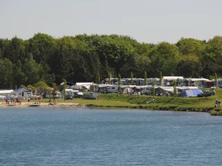 Am Ufer des Bernsteinsees stehen viele Zelte und Wohnwagen, umgeben von Bäumen und grünem Rasen.