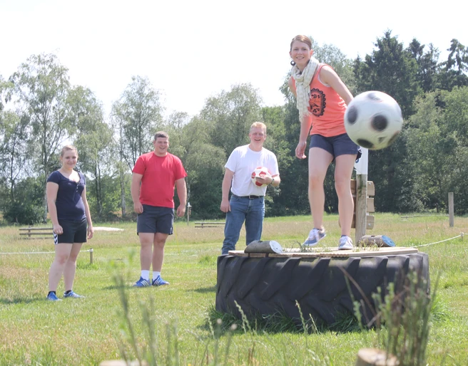 Fußballgolf Auf einer grünen Wiese spielt eine Gruppe von vier Menschen Fußballgolf. Eine Frau steht lächelnd auf einem großen Traktorreifen und kickt einen Fußball. Im Hintergrund lachen drei weitere Personen. Der sonnige Tag und die entspannte, spielerische Atmosphäre sind spürbar.