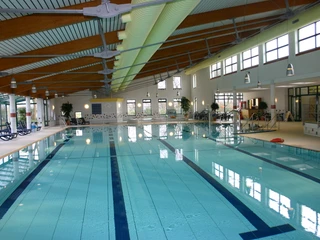 Hallenbad  Das Bild zeigt ein modernes Hallenbad mit einem großen Schwimmbecken, umgeben von hellen Fliesen und großen Fenstern, die natürliches Licht einlassen.