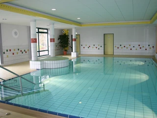 Bewegungsbecken Hallenbad Ein helles Hallenbad mit türkisfarbenem, gefliestem Bewegungsbecken, umgeben von großen Fenstern.